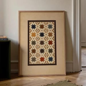 Puede incluir: Una obra de arte enmarcada con un diseño de colcha de estrellas. La colcha tiene un fondo beige con un patrón repetido de estrellas marrones, azules, doradas y rojas. El marco es de color madera clara y la obra está apoyada contra una pared blanca.