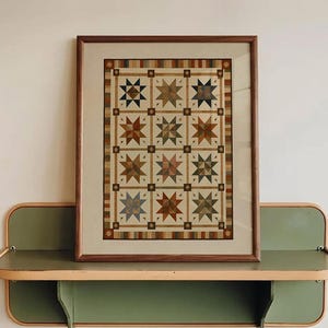 Può includere: Una stampa incorniciata con motivo a trapunta che presenta una griglia di dodici stelle multicolori nelle tonalità del blu, marrone e verde, inserita in una sottile cornice di legno, appoggiata su una mensola a muro in metallo verde salvia.