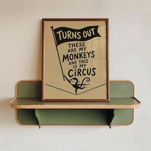 Op de afbeelding: Een ingelijste rechthoekige print met een beige achtergrond en zwarte tekst: TURNS OUT THESE ARE MY MONKEYS AND THIS IS MY CIRCUS. De print bevat een zwarte illustratie van een aap die een vlag vasthoudt terwijl hij op een koord balanceert.
