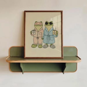 Peut inclure: Une impression d'art encadrée représentant deux grenouilles vertes en pyjama et peignoir. L'une porte un masque de sommeil rose et tient une tasse marquée COFFEE FIRST. L'autre porte des lunettes de soleil et un peignoir bleu, tenant une tasse GO WITH THE FLOW.