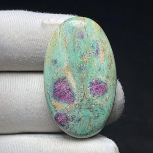 Puede incluir: Gema ovalada con una base de color turquesa e inclusiones magenta vibrantes. La superficie pulida muestra patrones naturales y variaciones de color. La gema está sobre un fondo de tela blanca. Dimensiones no visibles.