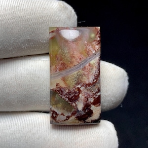 Op de afbeelding: Een rechthoekige edelsteen met een uniek patroon van rode, witte, gele en bruine tinten. De steen heeft een gepolijst oppervlak en een natuurlijke, organische uitstraling. De edelsteen wordt vastgehouden door een witte stof.