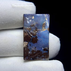 May include: A rectangular gemstone with a mix of blue, brown, and gold colors. The stone has a polished surface with a unique pattern of veins and textures. The gemstone is held by a gloved hand.