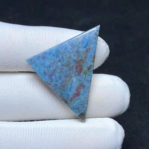 May include: A triangular-shaped polished gemstone featuring a mottled surface with shades of blue, deep red, and hints of green. The stone has a smooth, flat face with a distinct geometric shape and a natural, variegated mineral pattern throughout.