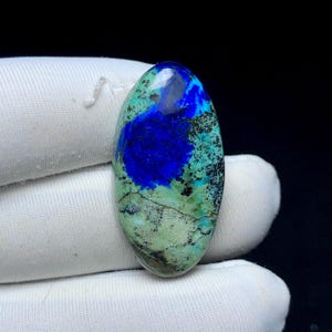 May include: Oval-shaped gemstone with a striking combination of colors. The stone features a deep blue central area surrounded by a mix of light green, turquoise, and black patterns. The polished surface reflects light, highlighting the natural textures and variations.