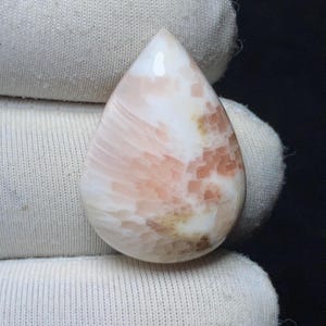 Könnte beinhalten: Ein tropfenförmiger Cabochon-Edelstein mit einer glatten, polierten Oberfläche und einer Mischung aus cremeweißen und sanften pfirsichfarbenen Mustern. Der Stein zeigt eine ausgeprägte radiale, fächerartige Textur mit feinen geometrischen Einschlüssen.
