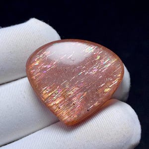 May include: A heart-shaped gemstone with a polished surface, exhibiting a warm orange-pink colour. The stone has iridescent flashes of colour, with streaks of yellow, pink, and green. The gem is held in a white glove, against a dark background.