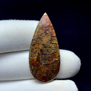 Puede incluir: Una gema pulida en forma de lágrima. La piedra presenta una mezcla de colores, incluyendo tonos marrones, naranjas y amarillos, con patrones más oscuros y moteados. La gema se encuentra sobre un fondo de tela blanca.