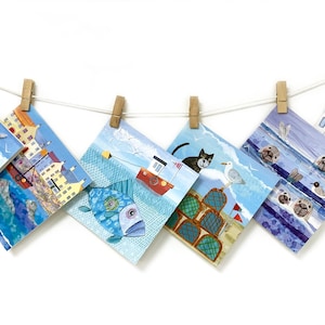 May include: A collection of square art prints featuring coastal illustrations, such as a fisherman, a red boat, a black cat, and seals. The prints are suspended from a string with wooden pegs against a white backdrop.