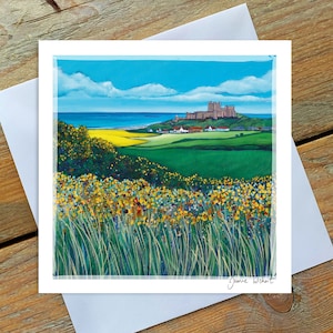May include: A painting depicting a castle perched on a hill overlooking a sandy beach with blue sea and white clouds. The castle is brown with a green roof. The hill is covered in green grass and yellow wildflowers. The painting is signed by the artist, Jeanne Wishart.