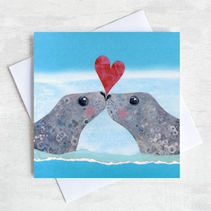 May include: A greeting card featuring a pair of grey seals with their noses touching, a red heart floating above them. The background is a light blue sky and water.