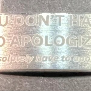 May include: A silver-colored bracelet with engraved text. The bracelet reads "YOU DON'T HAVE TO APOLOGIZE" in capital letters, with the smaller text "(you absolutely have to apologize)" below. The bracelet is a simple, elegant design.