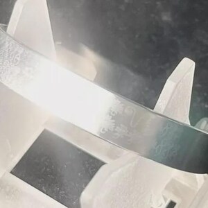 Könnte beinhalten: Ein silberfarbenes Armreif-Armband mit einem geätzten Blumendesign. Das Armband wird auf einem weißen Ständer vor dunklem Hintergrund präsentiert. Das Armreif ist ein schlichtes, elegantes Accessoire.
