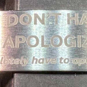 May include: A silver-colored metal cuff bracelet with the engraved text "YOU DON'T HAVE TO APOLOGIZE" and the text "(You absolutely have to apologize.)". The bracelet has a brushed metal finish.
