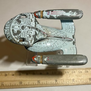 May include: A die-cast metal starship model featuring a light blue and grey hull with weathered paint, two elongated engine nacelles, and a saucer section. A wooden ruler is positioned below the model, showing a length of approximately 5.5 inches.