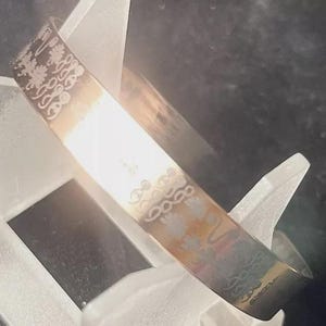 May include: A silver-toned cuff bracelet with etched floral and geometric patterns. The bracelet is displayed on a white stand inside a box. The design includes stylized flowers and repeating shapes, creating a delicate and intricate look.