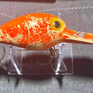 May include: A fishing lure with a bright orange and cream-colored mottled pattern. The lure features a prominent yellow eye with a black pupil, a long diving lip, and two silver metal hooks attached to the underside and rear of the body.