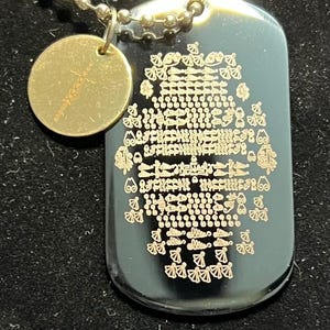 May include: A black rectangular dog tag pendant with a gold-toned metallic finish featuring an intricate, repeating etched pattern. Attached to the same silver-toned ball chain is a small, circular gold-toned charm engraved with a cross symbol.