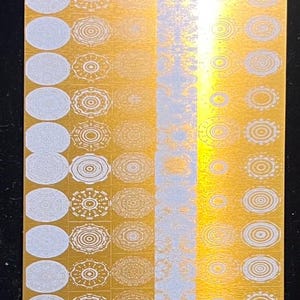 May include: A rectangular gold-coloured metal stamping plate featuring a grid of various circular patterns and designs. The surface displays multiple etched motifs, including concentric circles, floral-like shapes, and intricate geometric textures.