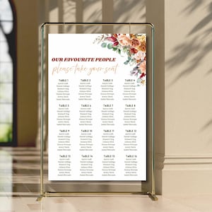 May include: A wedding seating chart displayed on a gold metal frame. The white paper features the text "OUR FAVOURITE PEOPLE please take your seat" in elegant script, with a floral arrangement in the upper right corner. The chart lists guest names under table numbers.