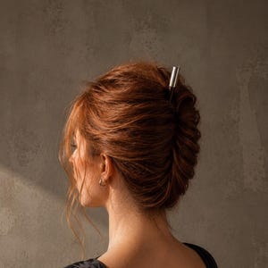 Può includere: Un bastoncino per capelli con un corpo in legno marrone scuro e una parte superiore in metallo argentato, inserito in un'acconciatura raccolta. L'accessorio è posizionato verticalmente tra i capelli, fissando le ciocche in una forma attorcigliata.