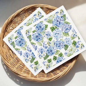 Può includere: Due tovaglioli di carta quadrati con un motivo floreale ad acquerello di ortensie blu e foglie verdi su sfondo bianco. I tovaglioli sono su un vassoio rotondo in vimini intrecciato.