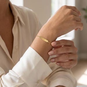 May include: A gold-colored bar bracelet. The rectangular bar is attached to a delicate chain. The bracelet is worn on a wrist, with a cream-colored long-sleeved shirt in the background.