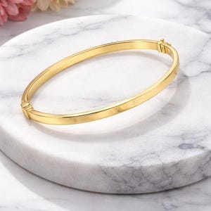 May include: A gold-colored bangle bracelet with a simple, elegant design. The bracelet is oval-shaped and has a clasp closure. The jewelry is displayed on a white marble surface, with pink and white flowers in the background.