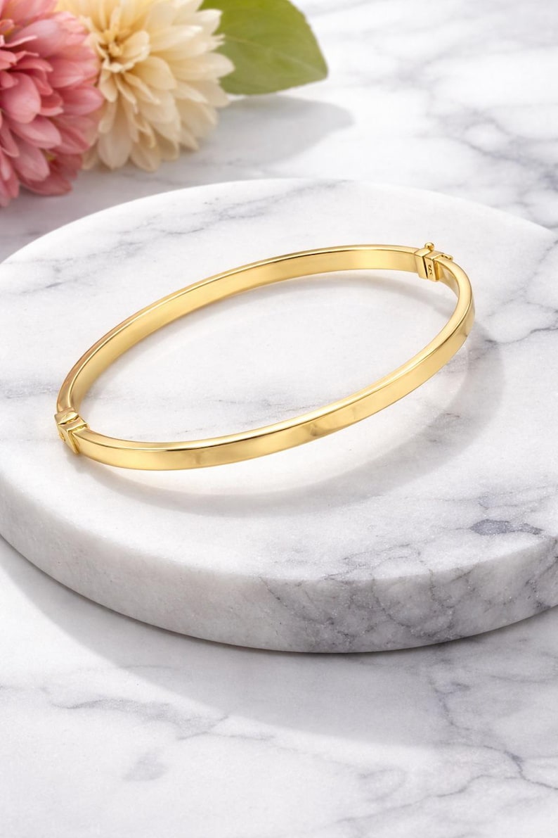 May include: A gold-colored bangle bracelet with a simple, elegant design. The bracelet is oval-shaped and has a clasp closure. The jewelry is displayed on a white marble surface, with pink and white flowers in the background.
