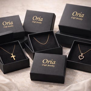 May include: Several black jewelry boxes with gold lettering that reads "Oria Craft Jewelry" are arranged on a satin surface. The boxes are open, revealing gold necklaces with a cross, a heart, and a simple chain.