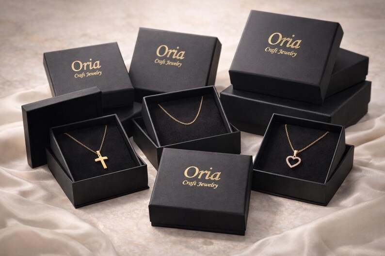 May include: Several black jewelry boxes with gold lettering that reads "Oria Craft Jewelry" are arranged on a satin surface. The boxes are open, revealing gold necklaces with a cross, a heart, and a simple chain.