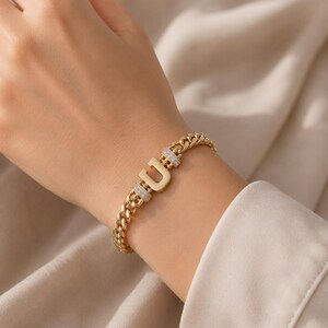 May include: A gold-toned chain link bracelet featuring a central letter U charm. The letter is flanked by two rectangular segments encrusted with small, clear, sparkling stones. The bracelet has a polished finish and a secure, integrated clasp design.