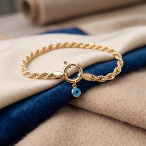 May include: A gold-colored rope chain bracelet with a circular clasp and a small charm. The charm is a blue eye with a gold border. The bracelet is resting on a blue and beige fabric.