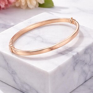 May include: A rose gold bangle bracelet with a simple, elegant design. The bracelet is displayed on a white marble block, with pink and yellow flowers in the background. The bracelet is approximately 2.5 inches in diameter.