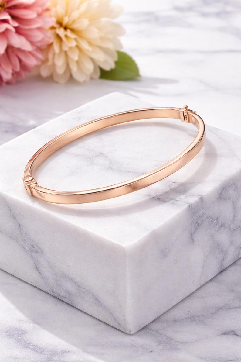 May include: A rose gold bangle bracelet with a simple, elegant design. The bracelet is displayed on a white marble block, with pink and yellow flowers in the background. The bracelet is approximately 2.5 inches in diameter.