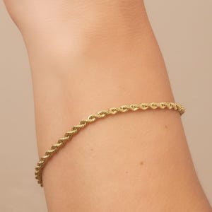 May include: A gold-colored rope chain bracelet. The bracelet is made of twisted metal links, creating a rope-like texture. The bracelet is worn on a person's arm.
