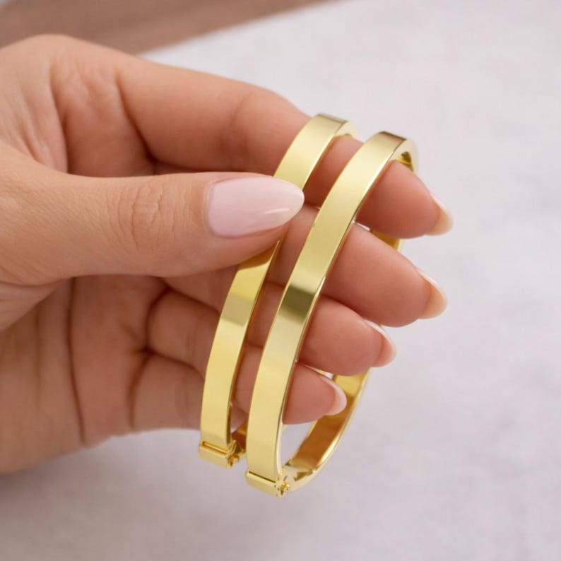 May include: Three gold-colored bangles are held in a hand. The bangles are rectangular in shape and have a smooth, polished surface. The bangles are approximately 2.5 inches in diameter. The background is a soft, neutral color.