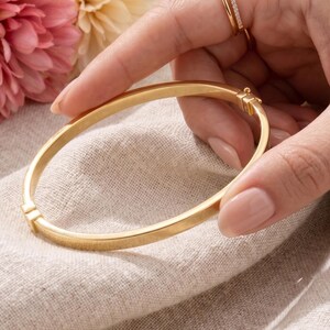 May include: A gold-colored bangle bracelet with a clasp is held in a hand. The bracelet is smooth and round. A second gold ring with small stones is on a finger. Pink and yellow flowers are in the background.