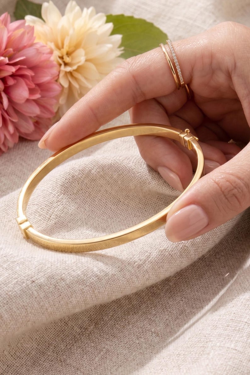 May include: A gold-colored bangle bracelet with a clasp is held in a hand. The bracelet is smooth and round. A second gold ring with small stones is on a finger. Pink and yellow flowers are in the background.