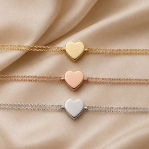 May include: Three delicate bracelets, each featuring a smooth, solid heart-shaped charm centered on a double-strand chain. The top bracelet is gold-toned, the middle is rose gold-toned, and the bottom bracelet is silver-toned in color.