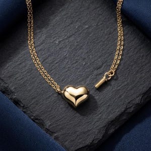 May include: A gold-colored heart pendant necklace. The heart-shaped pendant is smooth and reflective, suspended from a delicate double chain. The necklace is displayed on a dark, textured surface with a blue fabric accent.