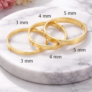 May include: Three gold-colored bangles of varying widths: 3 mm, 4 mm, and 5 mm. The bracelets are displayed on a white marble surface, with a soft-focus floral arrangement in the background. Each bangle has a clasp.