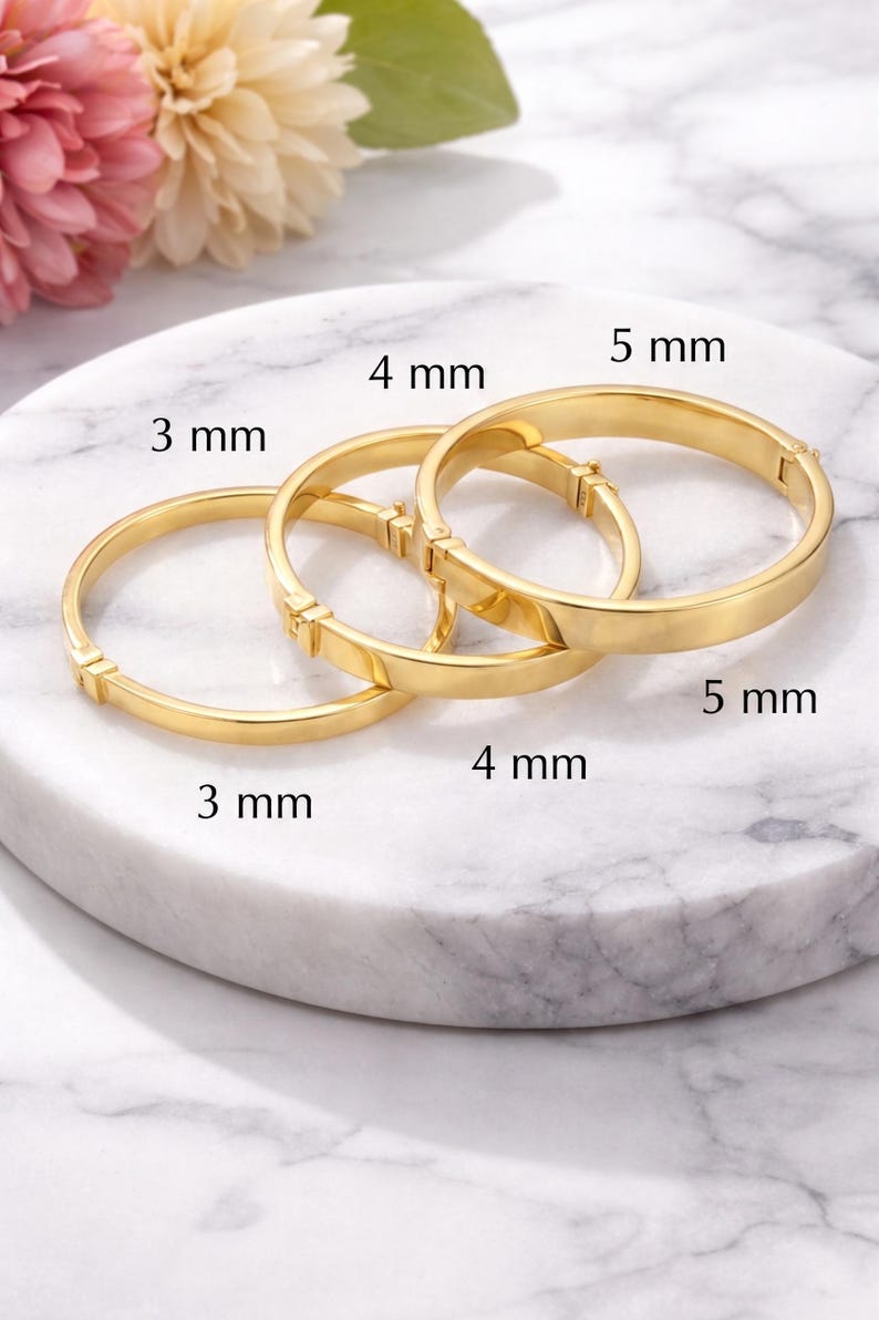 May include: Three gold-colored bangles of varying widths: 3 mm, 4 mm, and 5 mm. The bracelets are displayed on a white marble surface, with a soft-focus floral arrangement in the background. Each bangle has a clasp.