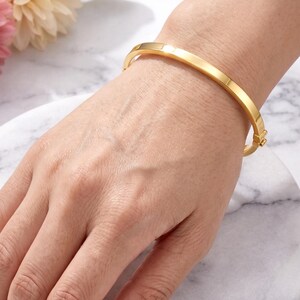 May include: A gold-colored, hinged bangle bracelet. The bracelet is a solid band with a rectangular profile. The clasp is visible on the side. The bracelet is worn on a wrist, with a marble background and flowers.