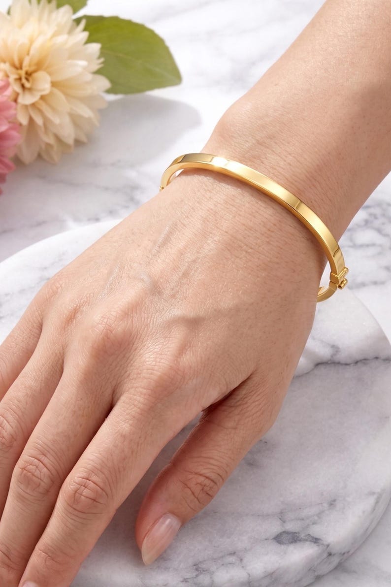 May include: A gold-colored, hinged bangle bracelet. The bracelet is a solid band with a rectangular profile. The clasp is visible on the side. The bracelet is worn on a wrist, with a marble background and flowers.