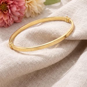 May include: A gold-colored bangle bracelet with a simple, elegant design. The bracelet is set against a neutral linen background, with pink and cream-colored flowers in the background. The bracelet appears to be made of metal.