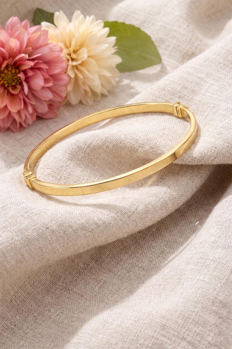 May include: A gold-colored bangle bracelet with a simple, elegant design. The bracelet is set against a neutral linen background, with pink and cream-colored flowers in the background. The bracelet appears to be made of metal.