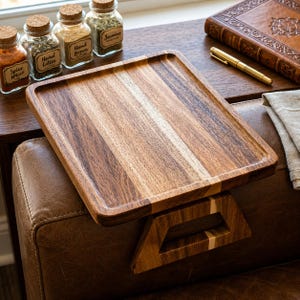 May include: A square wooden sofa tray with a dark brown finish and a light wood accent. The tray rests on a brown leather sofa. Small glass jars with cork lids and spice labels are visible in the background.
