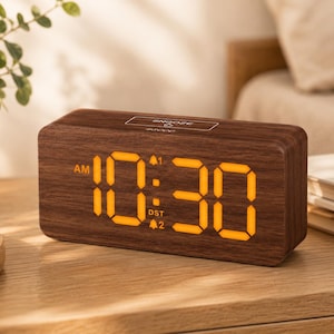 May include: A rectangular digital alarm clock with a dark brown wood grain finish. The front display features bright orange LED numbers showing 10:30, with AM, DST, and alarm indicators. A snooze button is located on the top surface of the clock.