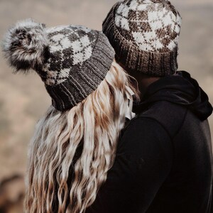 May include: Two matching gray and white argyle knit beanies. One beanie has a large faux fur pom-pom. The other person is wearing a black hoodie. The background is blurred, with a neutral color palette.